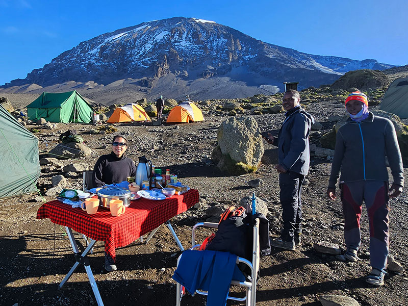 Trekkers enjoying breakfast at Karanga Camp on the Shira Route of Mount Kilimanjaro, with colorful tents and Kibo Peak in the background | Kili Quests