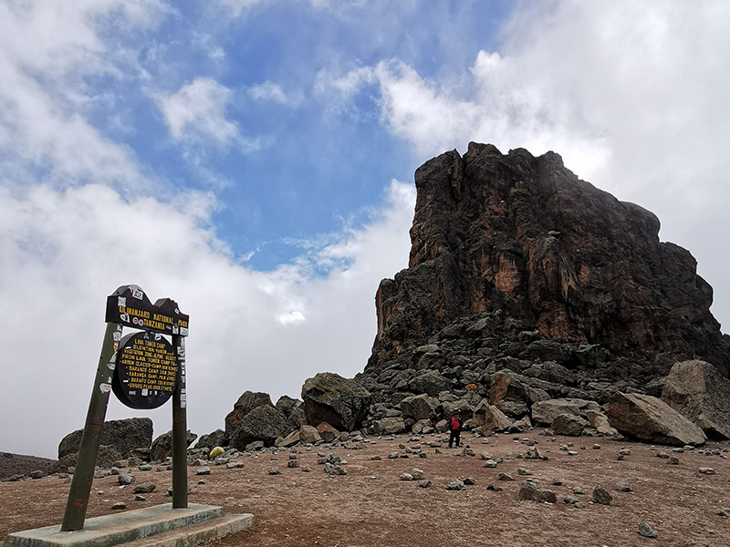 Lava Tower rising steeply at 4,630m on Mount Kilimanjaro’s Shira Route, with the trail sign and a trekker standing beneath the massive volcanic rock | Kili Quests