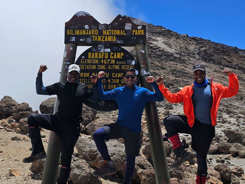 Three happy trekkers posing in front of the Barafu Camp sign on Kilimanjaro’s Northern Circuit Route, flexing muscles before summit day | Kili Quests