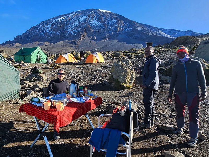 Trekkers enjoying breakfast at Karanga Camp on the Shira Route of Mount Kilimanjaro, with colorful tents and Kibo Peak in the background | Kili Quests