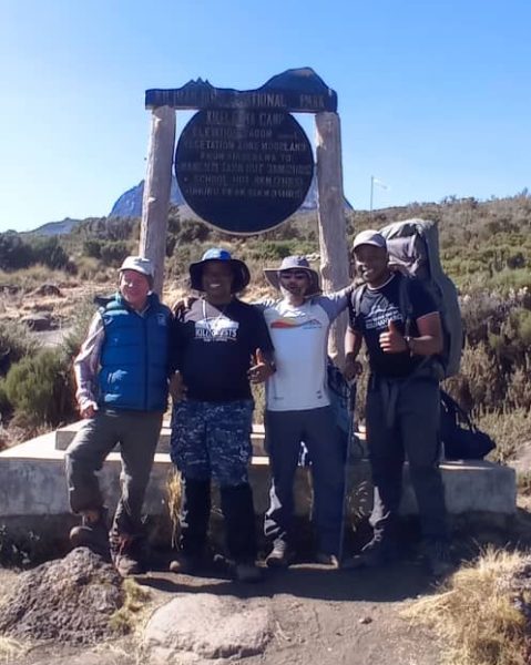 Group of Kilimanjaro trekkers posing at the Kikelelwa Camp sign on the Rongai Route, with Moorland vegetation and Mawenzi Peak in the distance | Kili Quests