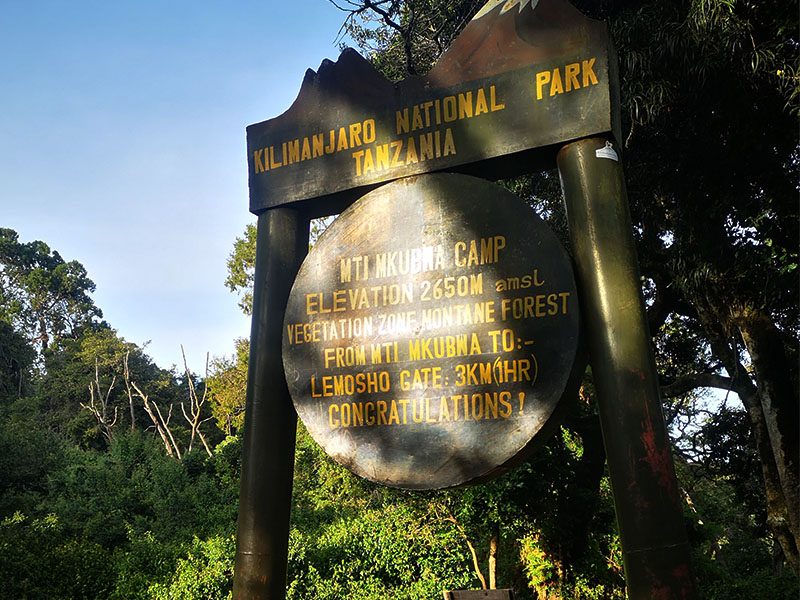 Mti Mkubwa Camp sign on Lemosho Route, Kilimanjaro National Park, showing elevation and distance to Lemosho Gate | Kili Quests