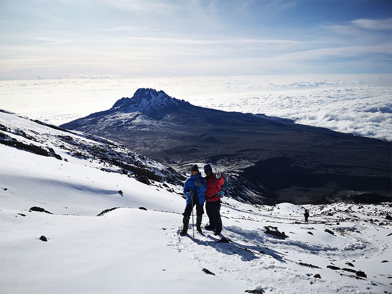 Two trekkers climbing through snow near Stella Point on Kilimanjaro’s Umbwe Route, with Mawenzi Peak and cloud-filled valleys below | Kili Quests