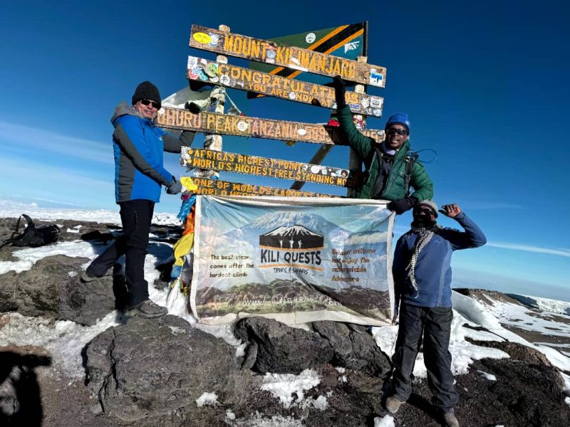 Smiling climber at Uhuru Peak holding a flag or sign after a successful Kilimanjaro ascent | Kili Quests