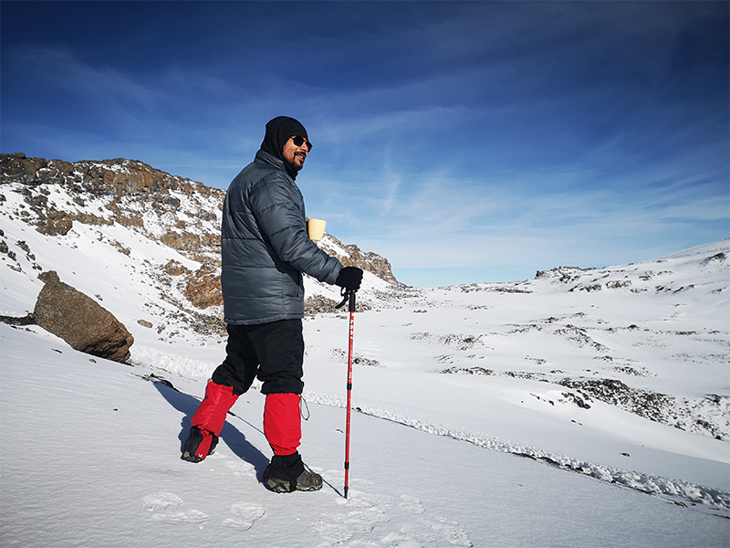 A climber stands on snow-covered terrain near Stella Point on Mount Kilimanjaro, holding a cup and trekking pole while overlooking the crater rim.