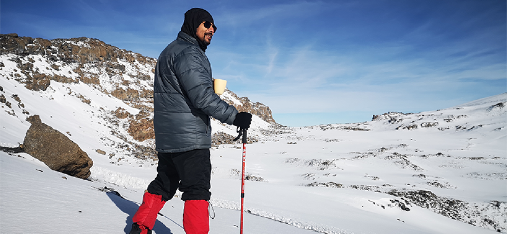 A climber stands on snow-covered terrain near Stella Point on Mount Kilimanjaro, holding a cup and trekking pole while overlooking the crater rim.